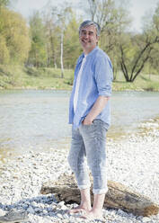 Happy man with hand in pocket standing by river - JBYF00113