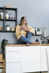 Nachdenkliche Frau mit Kaffeetasse auf dem Küchentresen sitzend - GIOF14816