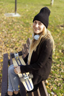 Lächelnde Frau mit Skateboard auf einer Bank sitzend - GIOF14847