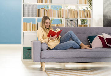 Junge Frau liest ein Buch auf dem Sofa zu Hause - GIOF14859