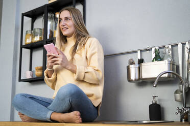 Nachdenkliche Frau mit Smartphone in der Küche - GIOF14862