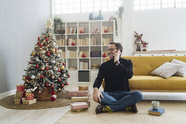 Glücklicher Mann, der am Weihnachtsbaum zu Hause mit seinem Handy telefoniert - GIOF14866