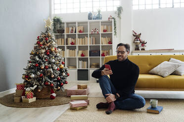 Mann mit Smartphone sitzt am Weihnachtsbaum im Wohnzimmer - GIOF14867