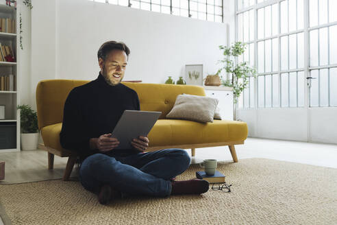 Älterer Mann sitzt auf einem Teppich und benutzt einen Tablet-PC zu Hause - GIOF14873