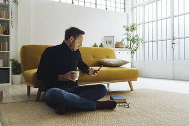 Älterer Mann mit Kaffeetasse und Smartphone im Wohnzimmer zu Hause - GIOF14874