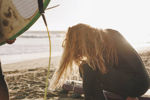 Frau mit langen Haaren hockt bei Sonnenuntergang am Strand neben einem Surfbrett, Gran Canaria, Kanarische Inseln - MRRF01879