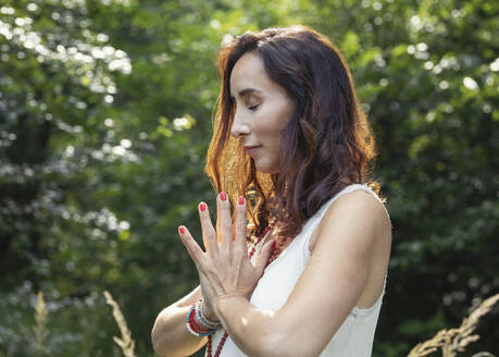 Frau mit geschlossenen Augen und gefalteten Händen beim Meditieren in der Natur - JBYF00120