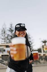 Smiling woman with beer cups standing at beach - MRRF01917