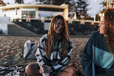 Fröhliche Frau hält Bierbecher und unterhält sich mit einem Freund am Strand - MRRF01926