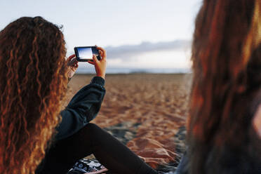 Junge Frau, die von einem Freund am Strand mit dem Smartphone fotografiert wird - MRRF01928