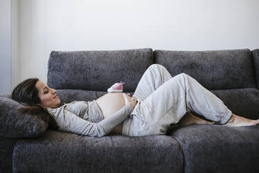Schwangere Frau mit Babyschuhen auf dem Bauch liegt zu Hause auf dem Sofa - EBBF05625