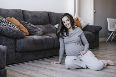 Smiling pregnant woman sitting by sofa at home - EBBF05633