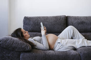 Pregnant woman using smart phone lying on sofa at home - EBBF05642