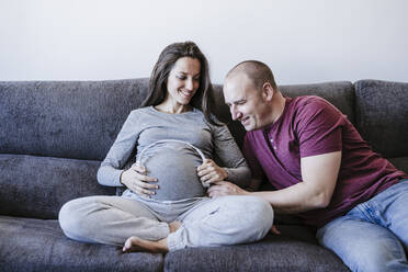Schwangere Frau hält Kopfhörer über den Bauch und schaut auf einen Mann, der auf dem Sofa sitzt - EBBF05649