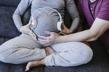 Pregnant woman with headphones over belly sitting with man at home - EBBF05650
