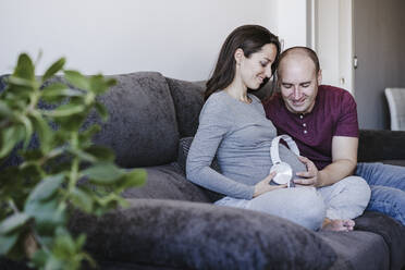 Glückliches Paar mit Kopfhörern über dem Bauch, das zu Hause auf dem Sofa sitzt - EBBF05651