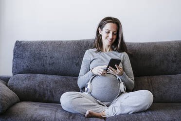 Schwangere Frau mit Kopfhörern über dem Bauch, die ein Smartphone benutzt, während sie zu Hause auf dem Sofa sitzt - EBBF05653