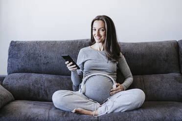 Pregnant woman holding smart phone with headphones over belly sitting on sofa at home - EBBF05654