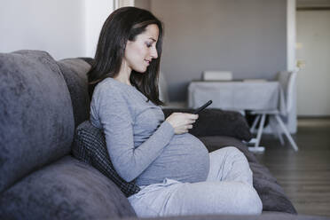 Pregnant woman using smart phone sitting on sofa at home - EBBF05655