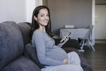 Smiling pregnant woman with smart phone sitting on sofa at home - EBBF05656