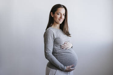 Pregnant woman holding belly standing in front of wall - EBBF05658