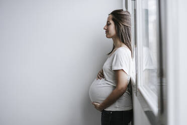 Pregnant woman leaning on window at home - EBBF05669