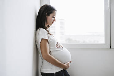 Pregnant woman leaning on wall near window at home - EBBF05671