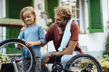 Smiling father looking at son cleaning bicycle outside house - JOSEF07064