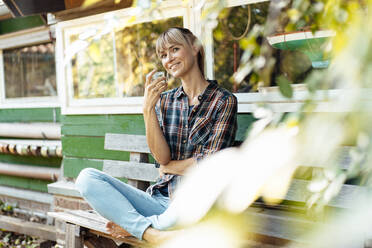Smiling woman with drinking glass on bench at garden - JOSEF07109
