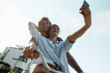 Happy couple on bicycle taking selfie through smart phone - JOSEF07166