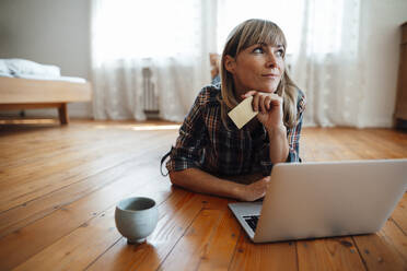 Thoughtful woman with credit card and laptop lying on floor at home - JOSEF07172