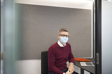 Businessman wearing protective face mask sitting on chair at office - JAHF00094