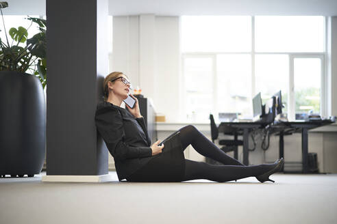 Geschäftsfrau, die auf dem Boden sitzend im Büro mit einem Smartphone spricht - JAHF00101