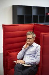 Nachdenklicher Geschäftsmann mit Laptop auf einem roten Stuhl im Büro - JAHF00134