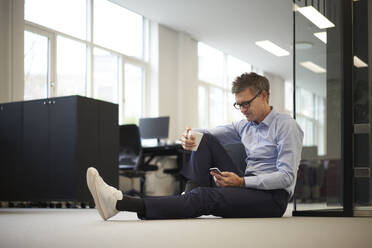 Businessman using smart phone sitting on ground in modern office - JAHF00139