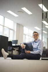 Smiling businessman with smart phone and coffee cup at office - JAHF00140