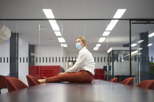 Businesswoman wearing protective face mask sitting on conference table in meeting room - JAHF00141