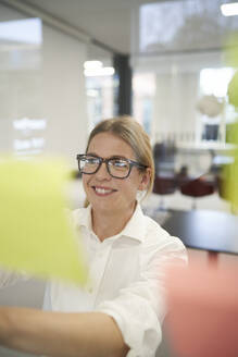 Happy businesswoman sticking adhesive note on glass wall in office - JAHF00164