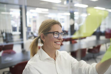 Smiling businesswoman looking at adhesive note on glass wall in office - JAHF00165