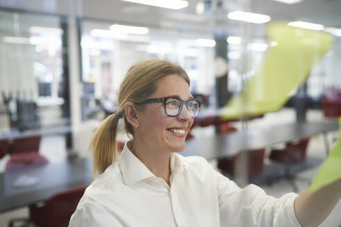 Smiling businesswoman looking at adhesive note on glass wall in office - JAHF00165