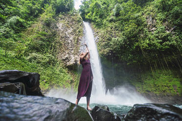 Junge Frau steht mit erhobenen Armen auf einem Felsen vor dem La Fortuna-Wasserfall, Costa Rica - RSGF00852