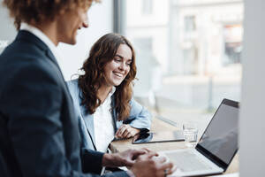 Happy businesswoman discussing with colleague using laptop in coworking office - GUSF06765