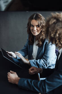 Smiling businesswoman with tablet PC looking at colleague sitting at desk in coworking office - GUSF06814