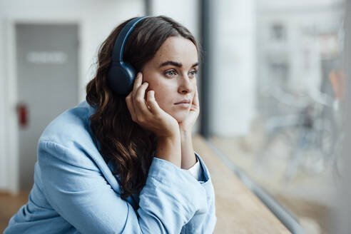 Nachdenkliche Geschäftsfrau mit Kopf in der Hand, die im Büro über drahtlose Kopfhörer Musik hört - GUSF06825