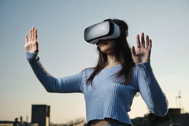 Young woman using virtual reality simulator gesturing in front of sunset sky - AGOF00231