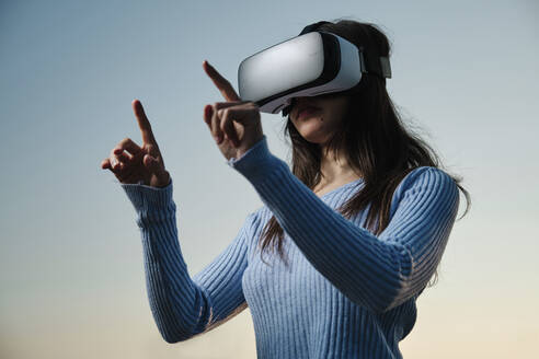 Junge Frau benutzt einen Virtual-Reality-Simulator und gestikuliert vor dem Sonnenuntergangshimmel - AGOF00232