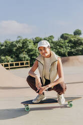 Smiling woman squatting on skateboard in park - JRVF02740