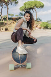 Smiling woman squatting with skateboard in park - JRVF02742