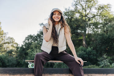 Smiling young woman sitting on skateboard in park - JRVF02746