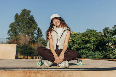 Lächelnde Frau sitzt auf einem Skateboard an einem sonnigen Tag - JRVF02747